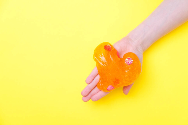 Orange slime in heart shape in kid hands. Girl hands playing slime toy on yellow background. Making slime. Copyspace. Love and valentines day concept - Valokuva, kuva