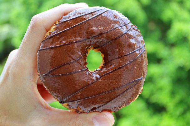 Mão segurando um donut revestido de chocolate contra desfocado Folhagem verde vibrante
 - Foto, Imagem