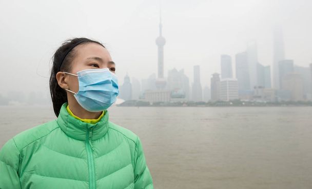 Worried Asian woman in green jacket and face mask looking away at city with pollution fog. - Valokuva, kuva