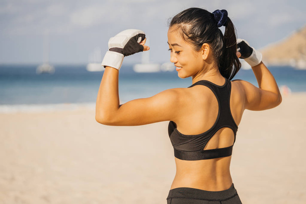 Athletic Asian woman in boxing gloves on the beach in summer shows biceps and smiles. Fighter, Training and Sport Lifestyle. - 写真・画像