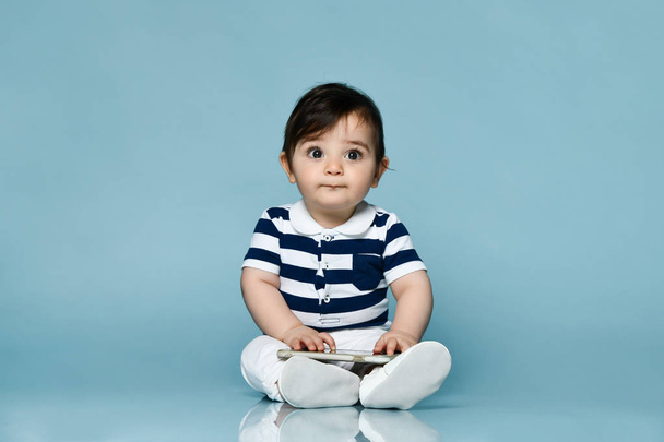 Toddler in striped t-shirt, white pants and booties. He holding smartphone, sitting on floor against blue background. Close up - Фото, зображення