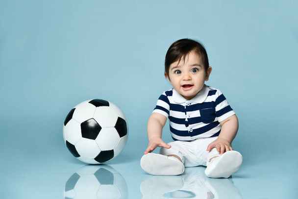 Toddler in striped t-shirt, white pants and booties. He is looking up, sitting on floor against blue background. Close up - Фото, зображення