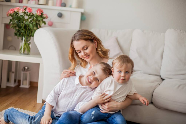 Hermosa madre joven, hijo pequeño, hija pequeña están sentados en el sofá en casa. Día de las Madres. Una familia feliz. Acogedor
. - Foto, Imagen