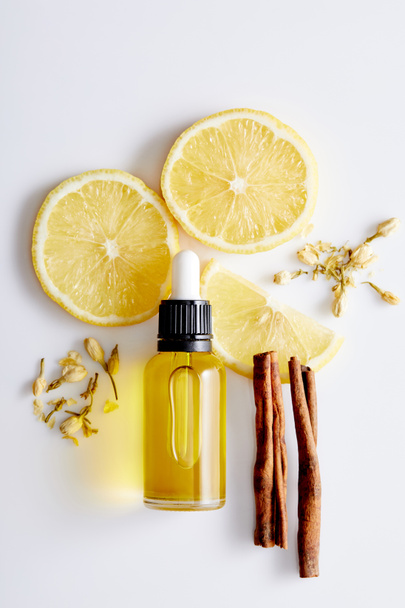 Top view of bottle of cosmetic oil with slices of lemon, sticks of cinnamon and vanilla buds on white background - Photo, Image