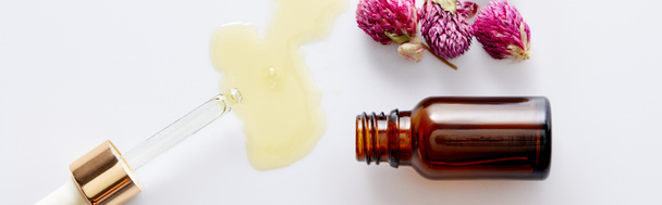 Horní pohled na kosmetický olej tekoucí z kapátka vedle láhve a růžové pupeny na bílém pozadí, panoramatický záběr - Fotografie, Obrázek