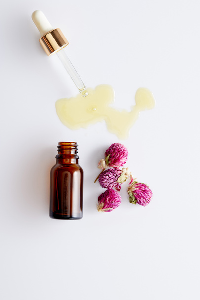 Widok z góry olejku kosmetycznego wypływającego z kroplomierza obok butelki i różowych pąków na białym tle - Zdjęcie, obraz