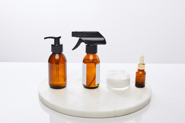 Dispenser bottles of oil and cosmetic cream on round stand isolated on grey  - Foto, imagen