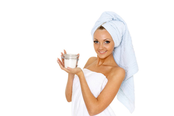 Jonge mooie vrouw verpakt in een handdoek, houdt een pot cosmetische hydraterende crème in handen op een witte achtergrond. Begrip lichaamsverzorging. - Foto, afbeelding