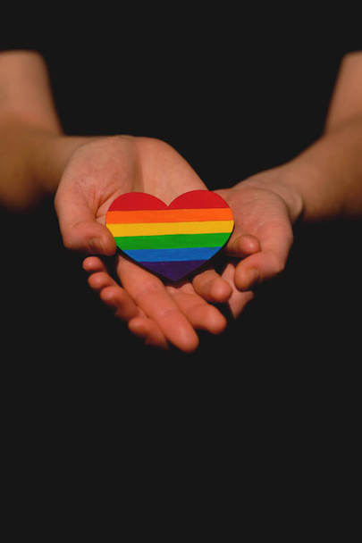 Женская рука, держащая сердце, окрашенное в цвета гордости ЛГБТ на тёмном фоне. Концепция Международного дня борьбы с гомофобией, сексуальное равенство, феминизм, социальная безопасность
 - Фото, изображение