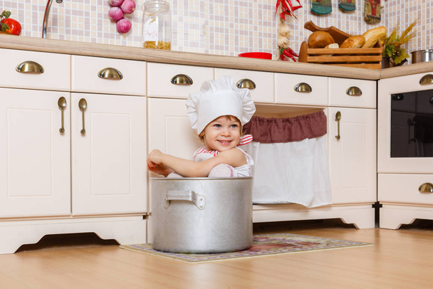 Little girl in apron and cap of the cook sitting  in the kitchen in the house. Mother's helper. 2 year old. - Foto, Bild