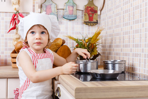 Little girl in apron and cap of the cook stands in the kitchen near cooker in the house. Mother's helper. 2 year old. - Foto, imagen