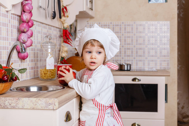 Little girl in apron and cap of the cook with an apple stands in the kitchen near sink at home. Mother's helper. 2 year old. - Photo, image