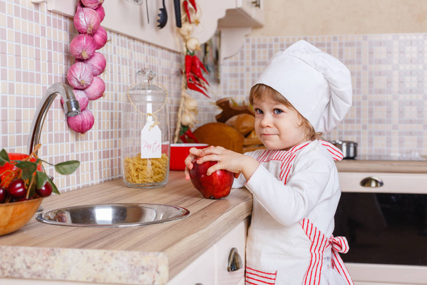 Little girl in apron and cap of the cook with an apple stands in the kitchen near sink at home. Mother's helper. 2 year old. - Zdjęcie, obraz