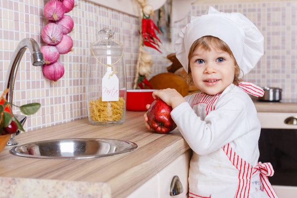 Niña en delantal y gorra del cocinero con una manzana se para en la cocina cerca del fregadero en casa. Ayudante de madre. 2 años de edad
. - Foto, imagen