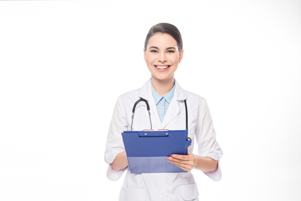 Mooie arts glimlachen op camera tijdens het schrijven op klembord geïsoleerd op wit - Foto, afbeelding
