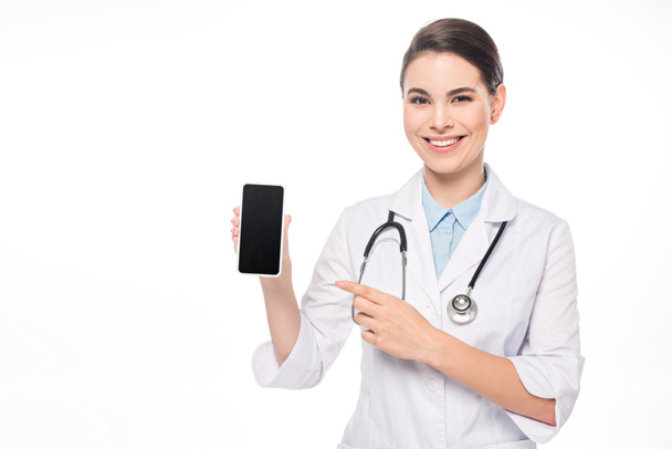 Attractive smiling doctor pointing at smartphone isolated on white - Photo, Image
