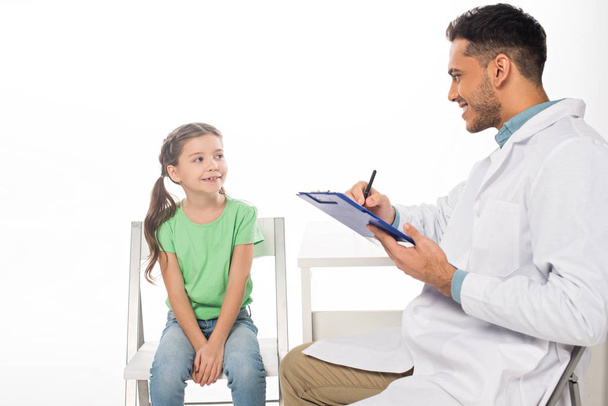 Smiling pediatrician looking at kid and writing on clipboard isolated on white - Fotoğraf, Görsel