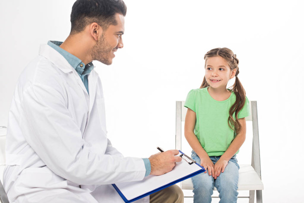 Smiling pediatrician with clipboard looking at kid on chair isolated on white - Foto, afbeelding