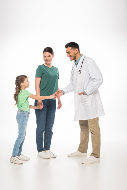 volle Länge des lächelnden Kinderarztes beim Händeschütteln mit Kind nahe der Mutter auf weißem Hintergrund - Foto, Bild