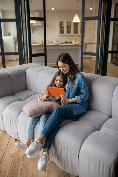 Daughter with tablet in her hands and mom sitting on couch. - Φωτογραφία, εικόνα