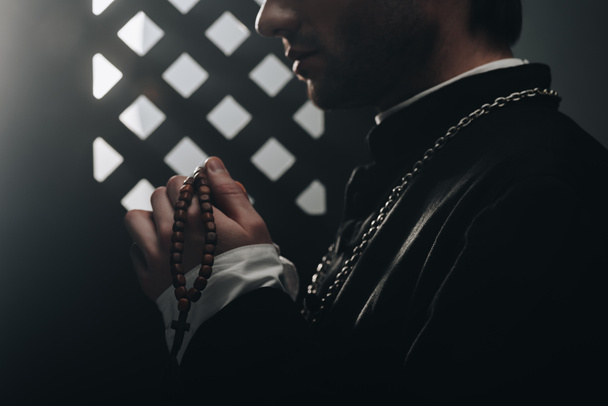 vista parcial del sacerdote católico sosteniendo cuentas de rosario de madera cerca de la rejilla confesional en la oscuridad con rayos de luz
 - Foto, Imagen
