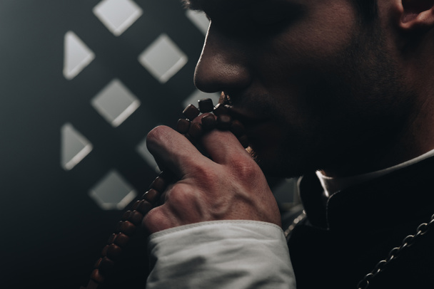 young catholic priest kissing wooden rosary beads near confessional grille in dark with rays of light - Fotoğraf, Görsel
