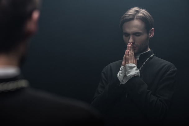 young serious catholic priest praying with closed eyes near own reflection isolated on black - Foto, imagen