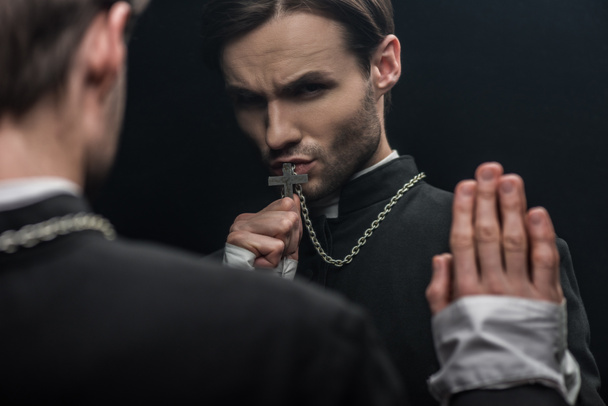 young tense catholic priest kissing holy cross while looking at own reflection isolated on black - 写真・画像