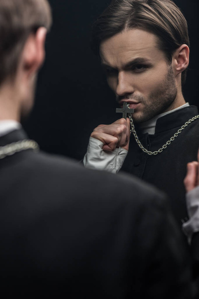 young tense catholic priest kissing holy cross while looking at own reflection isolated on black - Photo, Image