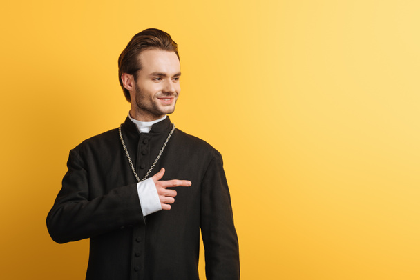 smiling catholic priest looking away and pointing with finger isolated on yellow - Фото, изображение