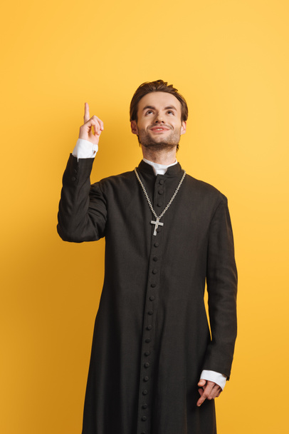 усміхнений католицький священик дивиться вгору і вказує пальцем ізольовано на жовтому
 - Фото, зображення