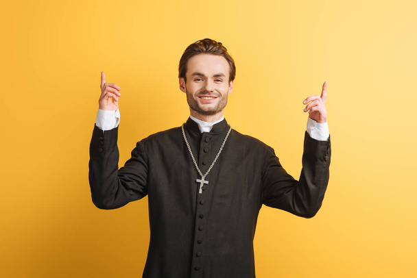 Lächelnder katholischer Priester zeigt mit den Fingern nach oben, während er isoliert auf gelb in die Kamera schaut - Foto, Bild