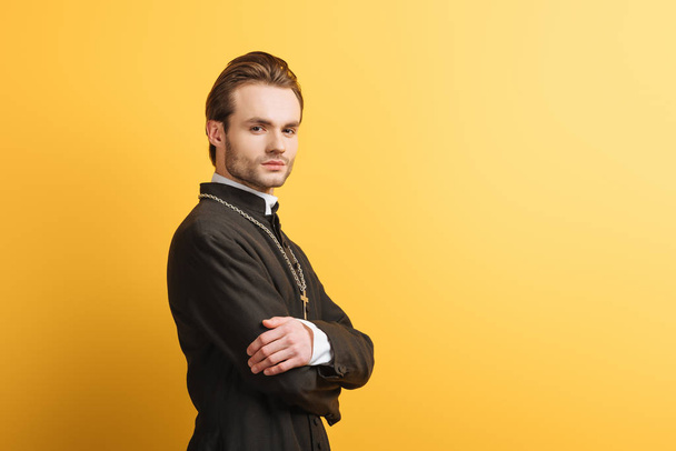 confident catholic priest with crossed arms looking at camera isolated on yellow - Фото, изображение