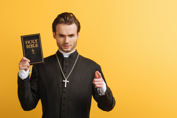 serious catholic priest pointing with finger at camera while holding bible isolated on yellow - Foto, afbeelding