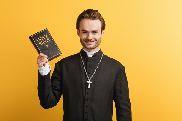 happy catholic priest holding bible while smiling at camera isolated on yellow - Fotoğraf, Görsel