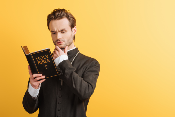 soustředěný katolický kněz dotýkající se obličeje při čtení bible izolované na žluté - Fotografie, Obrázek