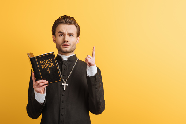 thoughtful catholic priest looking up and showing idea sign while holding open bible isolated on yellow - Fotoğraf, Görsel