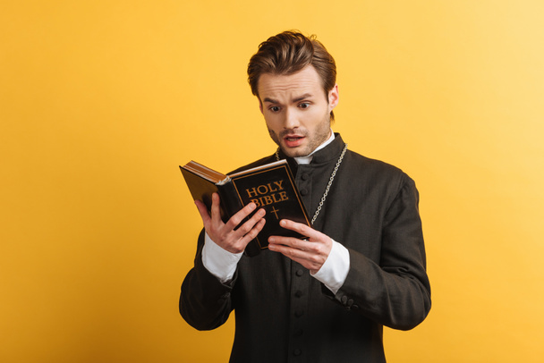 shocked catholic priest reading holy bible isolated on yellow - Foto, Imagen
