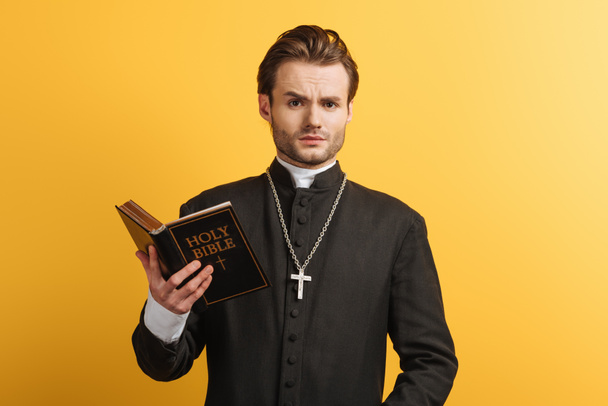 surprised catholic priest looking at camera while holding bible isolated on yellow - Fotografie, Obrázek
