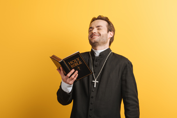 Sacerdote cattolico eccitato che ride mentre tiene la santa bibbia isolata sul giallo
 - Foto, immagini