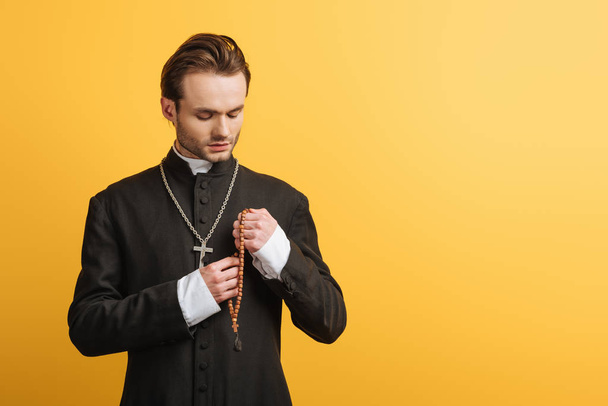 young thoughtful catholic priest holding wooden rosary beads isolated on yellow - Photo, Image