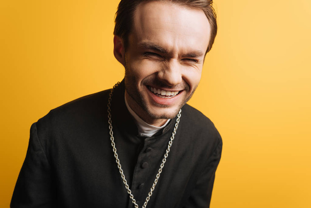 excited catholic priest laughing while looking at camera isolated on yellow - Fotó, kép