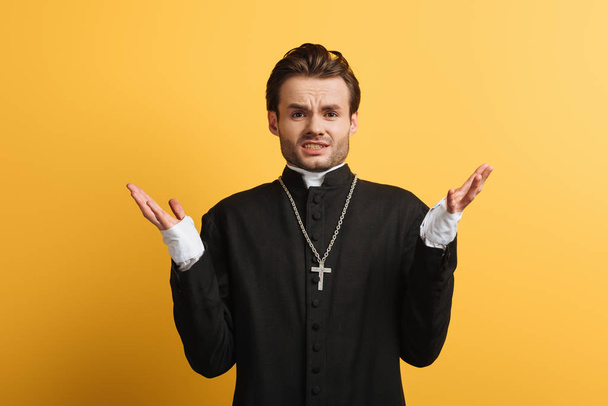 znechucený katolický kněz ukazující gesto pokrčení ramen před kamerou izolovanou na žluté - Fotografie, Obrázek