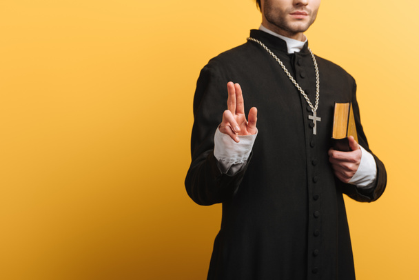 partial view of catholic priest showing blessing gesture while holding bible isolated on yellow - Fotoğraf, Görsel