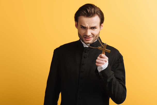 angry catholic priest showing cross while looking at camera isolated on yellow - Foto, afbeelding