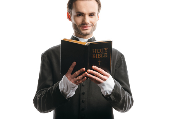 young, smiling catholic priest holding holy bible while looking at camera isolated on white - Fotoğraf, Görsel