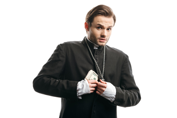 vyděšený katolický kněz při pohledu na kameru, zatímco skrývá peníze pod sutanou izolované na bílém - Fotografie, Obrázek