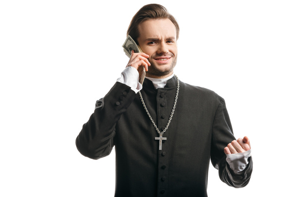 corrupt catholic priest looking at camera while holding money near head isolated on white - Foto, afbeelding