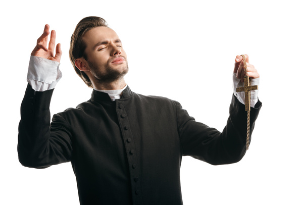 young catholic priest praying with closed eyes and raised hands while holding golden cross isolated on white - 写真・画像