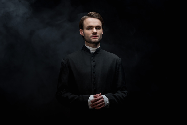 young serious catholic priest standing with folded hands on black background with smoke - Fotoğraf, Görsel
